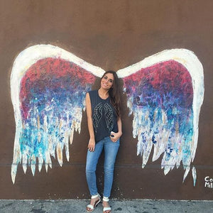 Grey tank with silver and black wings featured on a girl against a colorful mural of angel wings. 