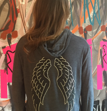 Load image into Gallery viewer, Girl wearing the grey hoodie with silver and black wings on the back. She is standing in front of a colorful wall.