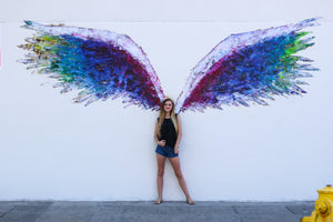 Black tank top with black wings, on the front, featured on a teenager girl standing in front of a mural with colorful angel wings. 