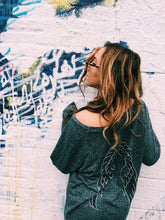 Load image into Gallery viewer, Cozy Comfy Crewneck (a grey shirt with black and silver wings on the back), worn on a girl against a white colorful wall.