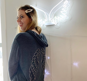 President & founder of the brand wearing the grey hoodie with silver and black wings on the back, standing in front of a white neon angel wing sign. 