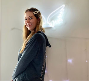 President & founder of the brand wearing the grey hoodie with silver and black wings on the back, standing in front of a white neon angel wing sign.