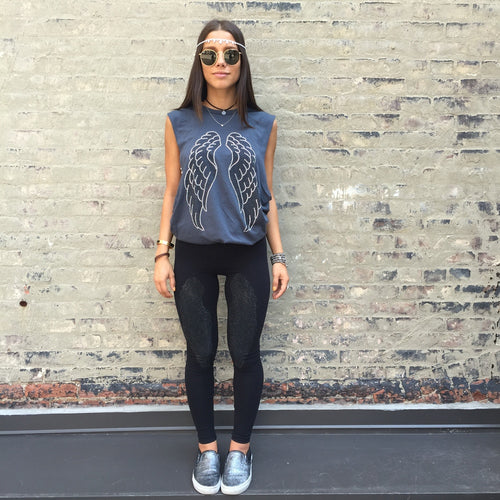 Model against a light brick wall wearing grey tank with silver and black wings on it. 