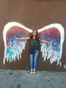 Girl wearing grey tank with silver and black wings against a colorful angel wing wall. 