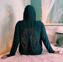 Load image into Gallery viewer, Back of grey hoodie with silver and black angel wings. The model is sitting on a bed with fairy lights around her. 