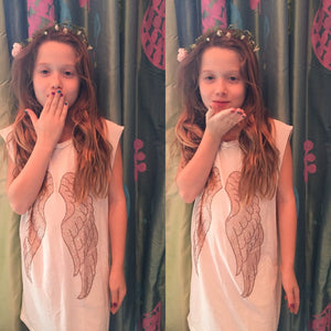 Photo of a little girl wearing a flower crown, and our Heart of Gold Tank. It is a white tank with rose gold wings. 